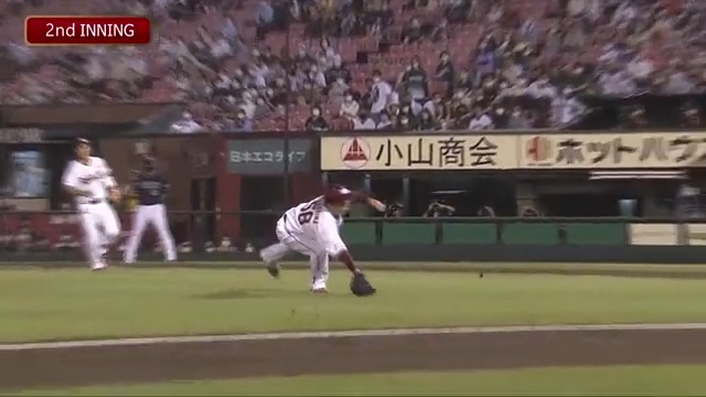 A baseball player in a white uniform makes a low-fielding catch near the foul line during the 2nd inning, with Japanese signage for "小山商会" and "ホットパ" visible on the outfield wall, and the caption "何これ！かっこいい" ("What is this! So cool!") reflecting admiration for the impressive defensive play.