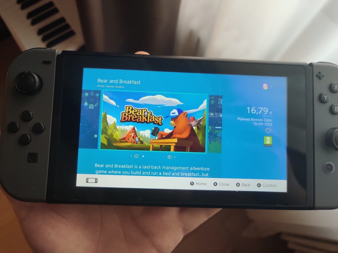 A photo of a hand holding a gray Nintendo Switch. On it, we see the eShop page for Bear and Breakfast. In the background, a slightly messy floor with what could be construed as a blurry toe. Didn't know why I felt the need to add that last part.