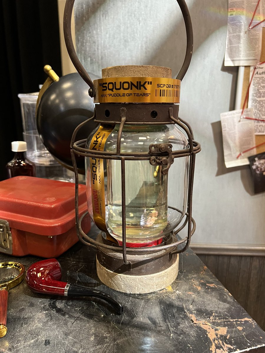 A photo of the same office, but the focus is on the captured squonk. Said squonk is trapped in a portable hand lantern, and the label towards the top of it reads "SQUONK" in a bold font with capital letters. Below that, it says "AKA "PUDDLE OF TEARS"" in the same font at a different size. To the right, some more text reads "SCP DB #73177-", but the rest of it isn't visible from the camera's angle.