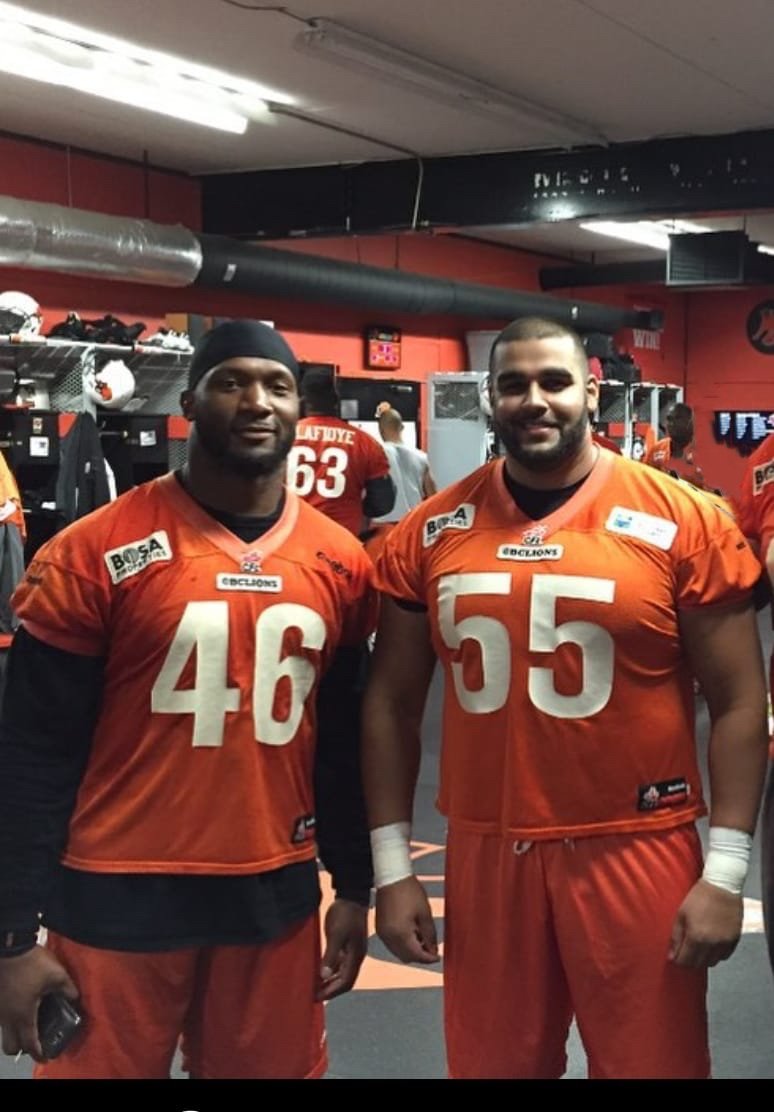 A photo of two large built men standing in a locker room. The locker room is busy and the walls are painted a bright orange. Both men are wearing orange BC Lions jerseys, numbered 46 and 55.