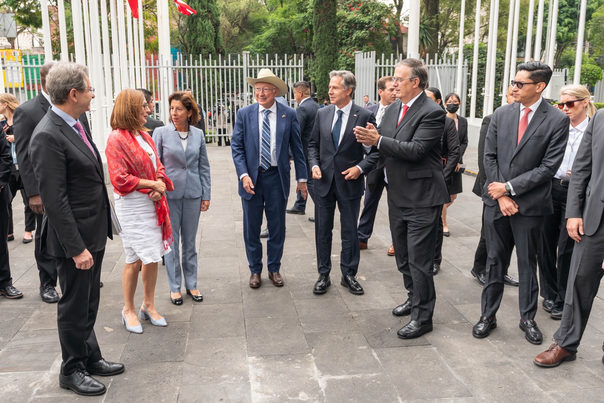 A group of U.S. and Mexican government leaders, including Secretary Blinken and U.S. Commerce Secretary Raimondo,  as well as Mexican Foreign Secretary Ebrard, stand in a close group talking in front of a metal fence in Mexico City.