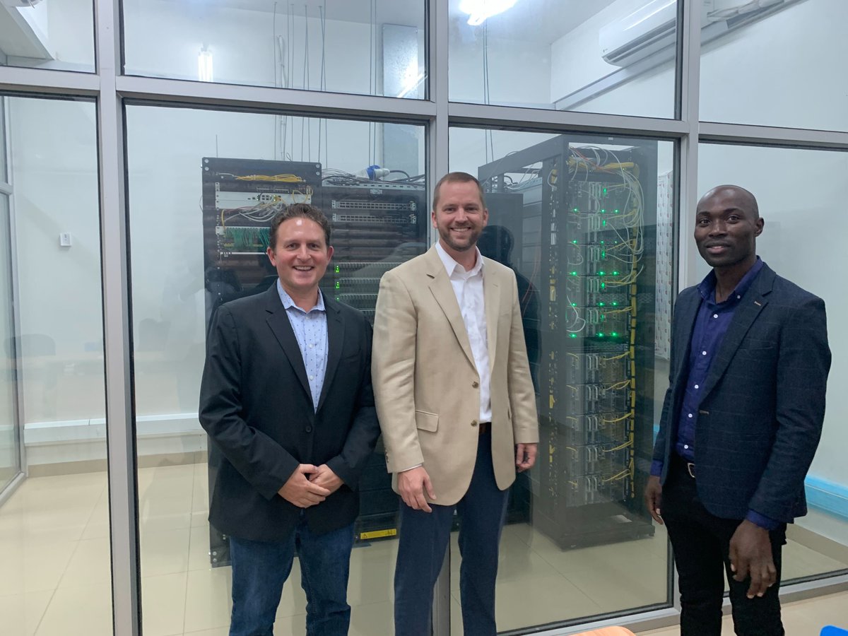 TACC's tweet photo. Dir. of Special Projects John Fonner (mid) represented TACC at the African Centers of Excellence in Bioinformatics and Data-intensive Sciences consortium meeting. Also pictured, Ari Berman, BioTeam (left); Rodgers Kimera, @AceUganda (right). Stampede1 nodes in back. @USAmbUganda https://t.co/polev8SJ86