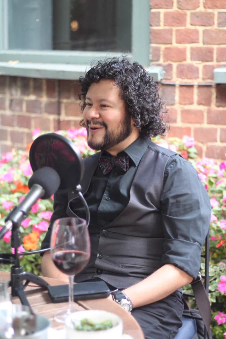 Arturo sentado fuera en una terraza de un bar. Está hablando en el micrófono y sonriendo.