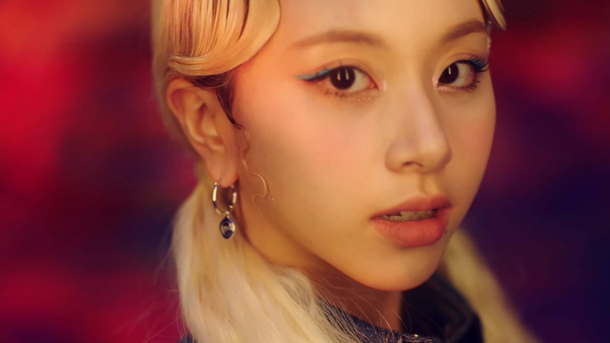 A close-up portrait of a blonde woman with blue eyeliner, red lipstick, and dangling earrings against a red background.