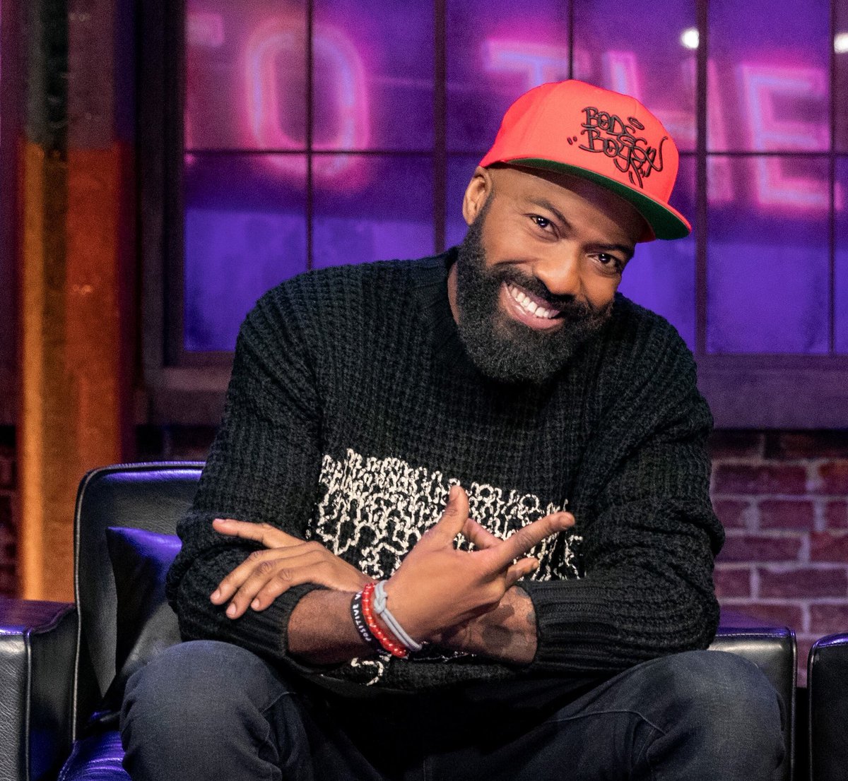 Desus smiling while sitting on the set of the Desus & Mero show. He's wearing an orange cap and a black sweater, with his elbows on his knees.