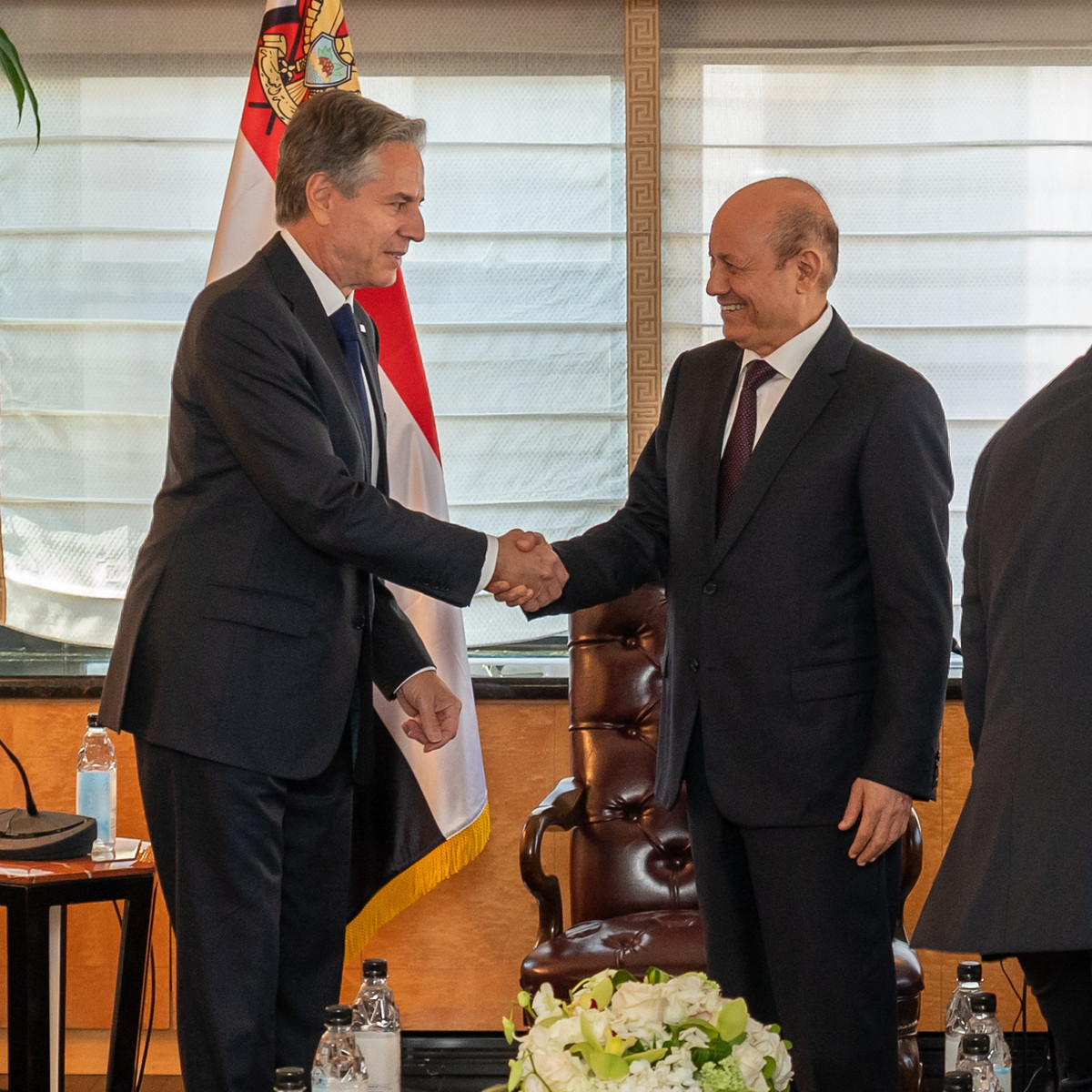 Secretary of State Antony Blinken stands with Yemeni Chairman of the Presidential Leadership Council Rashad Al-Alimi in front of windows with lowered blinds. The two men shake hands and smile in front of some chairs and flags.