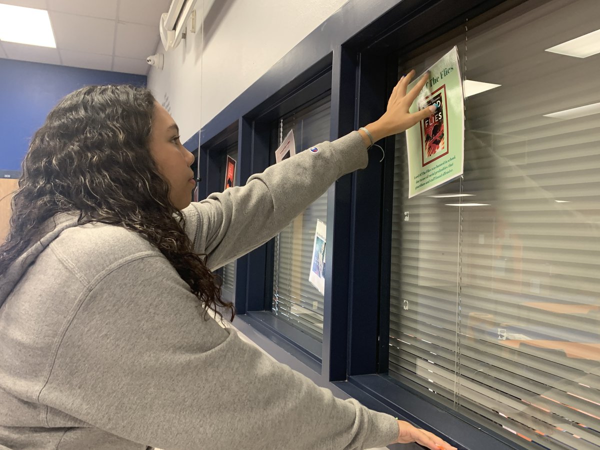 SehSeminoles's tweet photo. To celebrate Banned Book Week, Allison R. hangs banned book posters created by the Digital Media students to go along with the Banned Books display created by Ms. Rohde and the media aides in the Southeast High School Media Center. https://t.co/HZzHsgqLE3