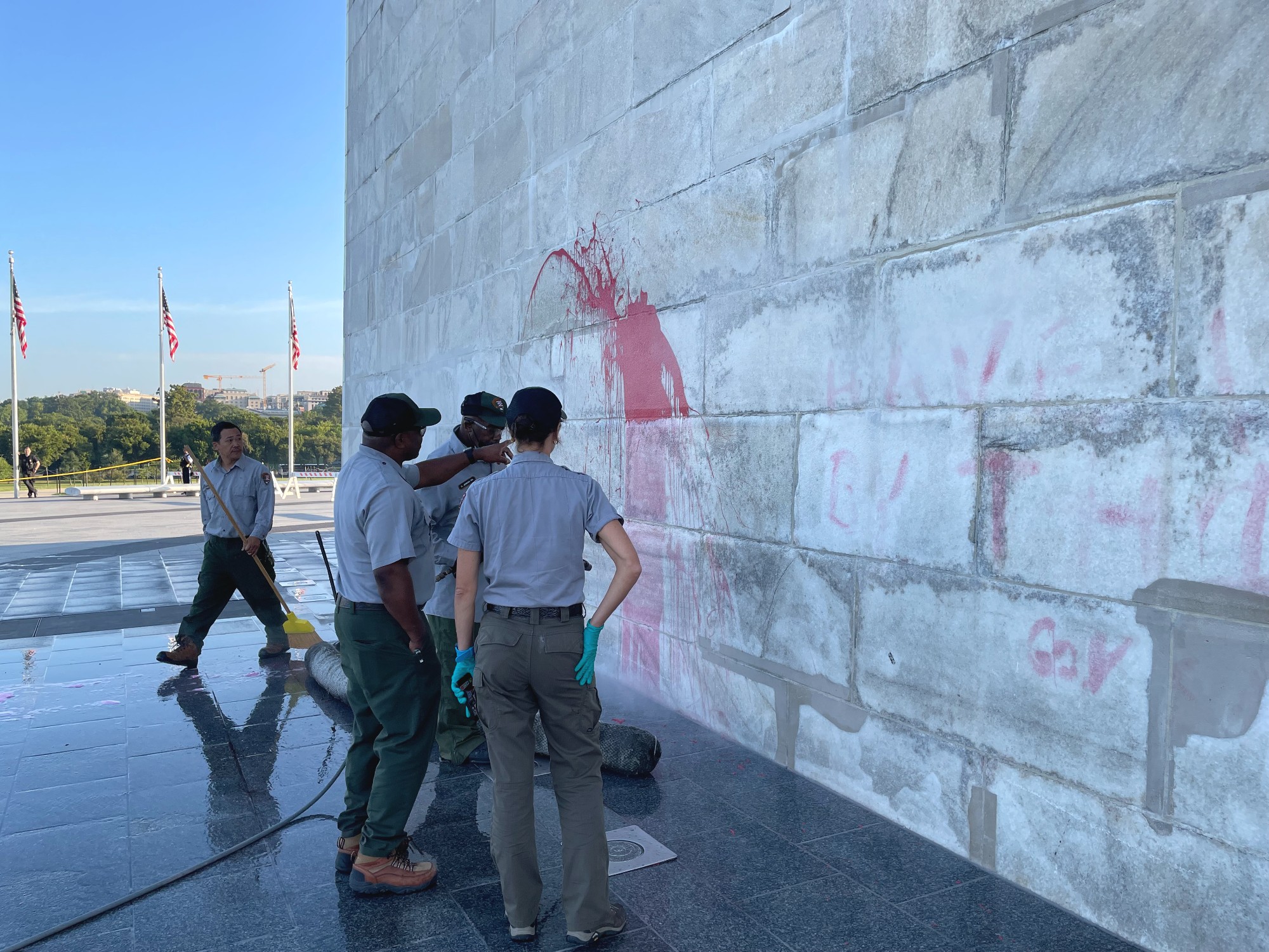 Four National Park Service employees stand next to the white stone base of the Washington Monument looking at red paint smeared on the wall.