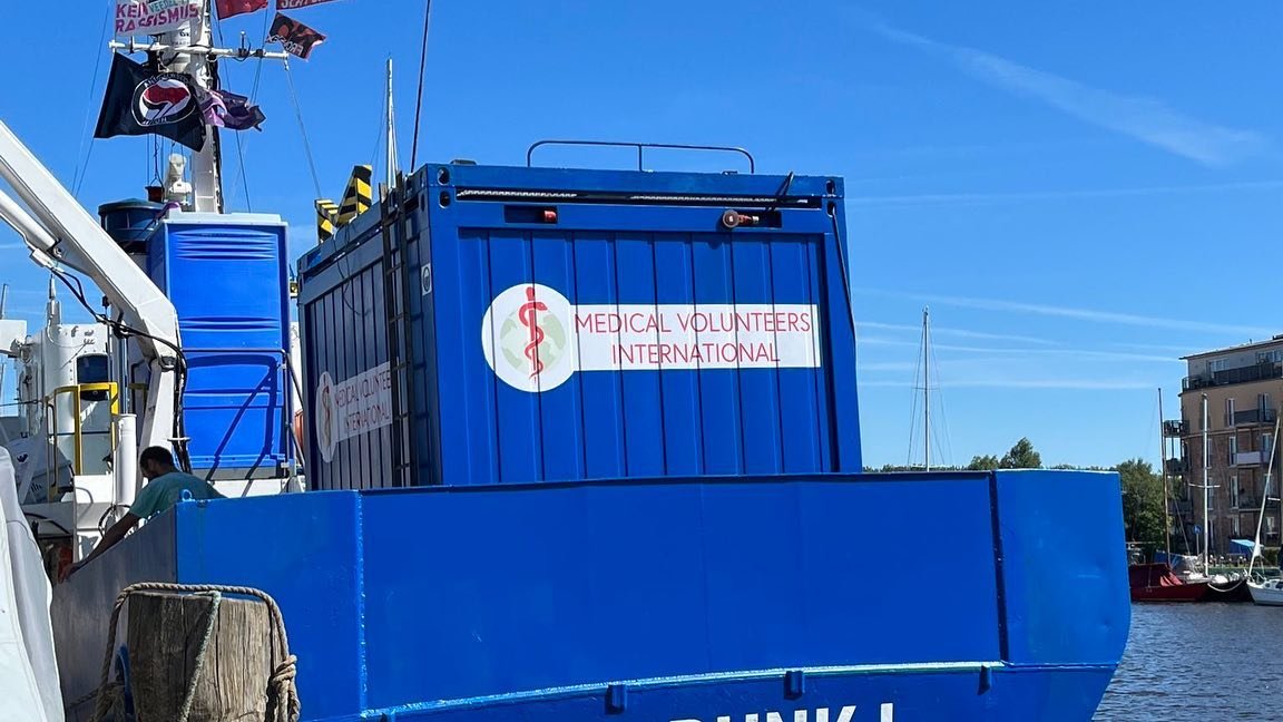 Auf dem Heck der SEA PUNK I ein blauer Container mit dem Logo u. Aufschrift von Medical Volunteer International. Daneben ein Dixiklo. AM Fahnenmast eine Antifa-Fahne, ein Banner mit der Aufschrift 'Kein Rassismus' und eine 'No Frontex'-Fahne.