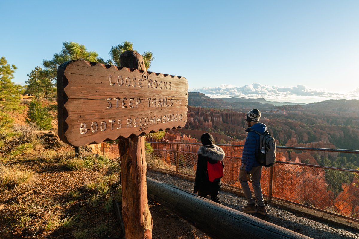 Wooden sign reading Loose rocks, Steep Trails, Boots Recommended. Two hikers step along path beside it. In the background a vast landscape of red rock spires and cliffs beneath a blue sky.