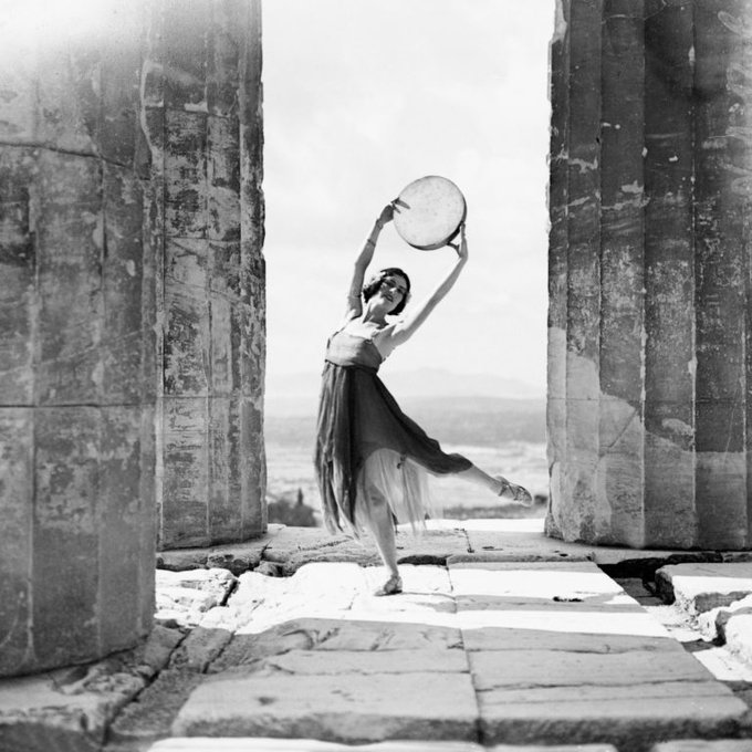 Monochrome photograph featuring a white woman posed as if dancing with one leg raised to the side and holding a circular disc above her head, wearing a dress with loose fabric, standing between large stone columns on stone slabs