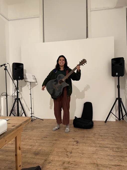 Ahana sings with an acoustic guitar in front of a white wall with black PA system behind.
