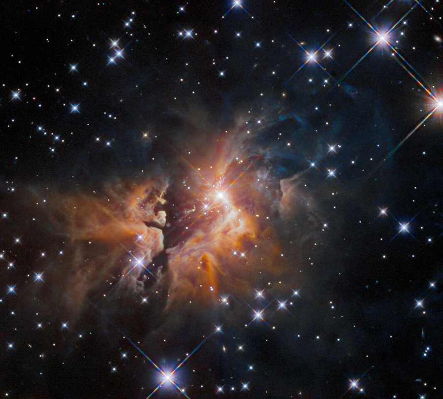 A glowing, young star at the center of the image is surrounded by thick shrouds of orange gas and dust. Towards the edges of the image, bright stars shine against a black backdrop of space.