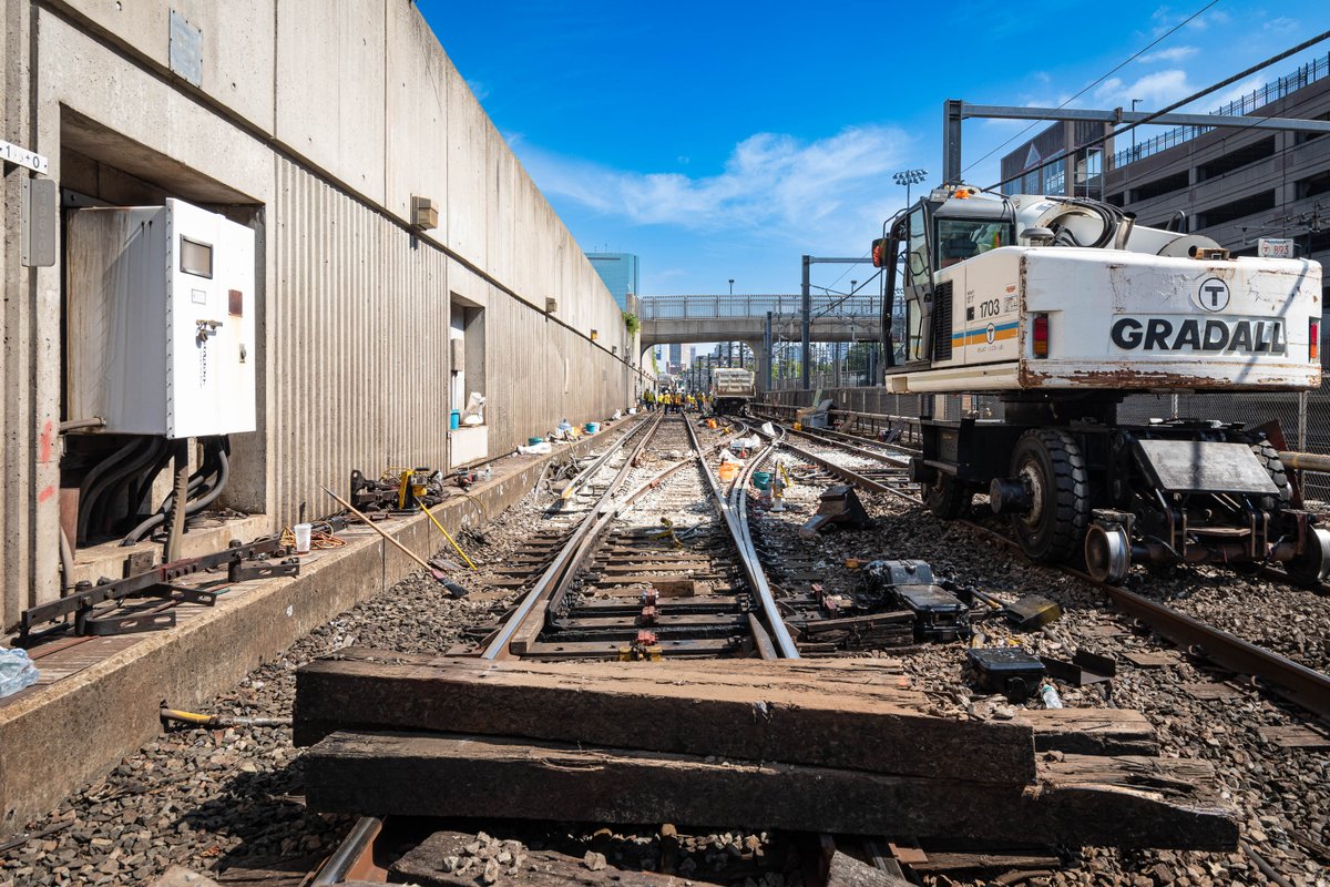 Old wooden rail ties stacked on top of the Orange Line rails outside of Ruggles with a rail-bound construction vehicle on the right, looking down at the crossover.