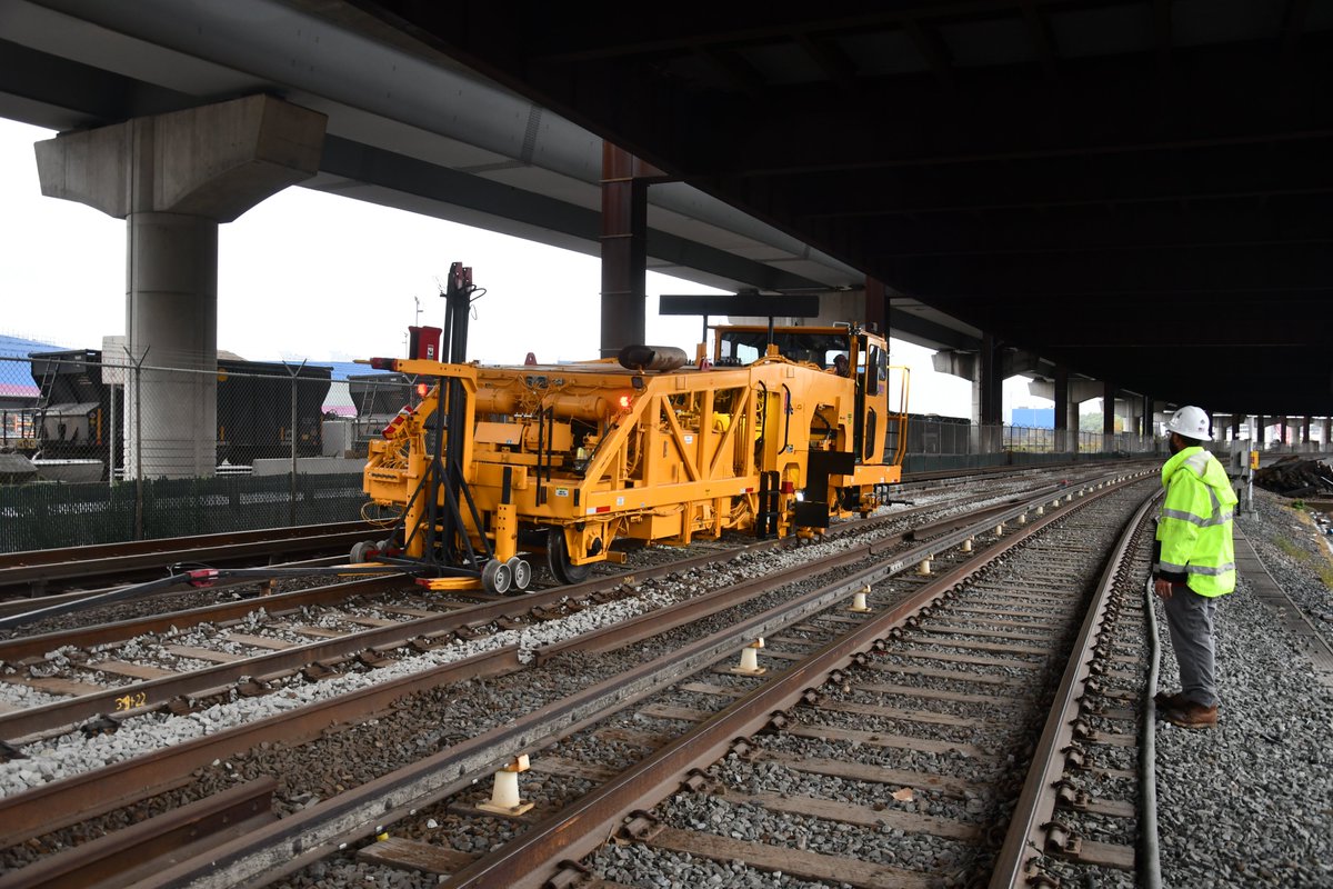 A yellow rail-bound tamping machine running along the Orange Line tracks outside of Community College under the I-93 interstate roadway above.