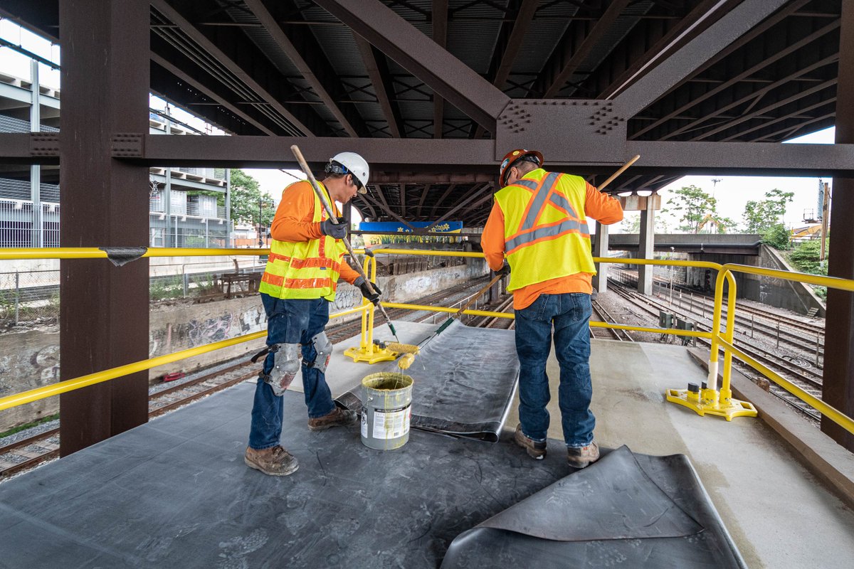 Two construction workers in yellow safety vests and hard hats using paint rollers to apply adhesive to the weatherproofing membranes on the roof of Sullivan Square station under the I-93 roadway.