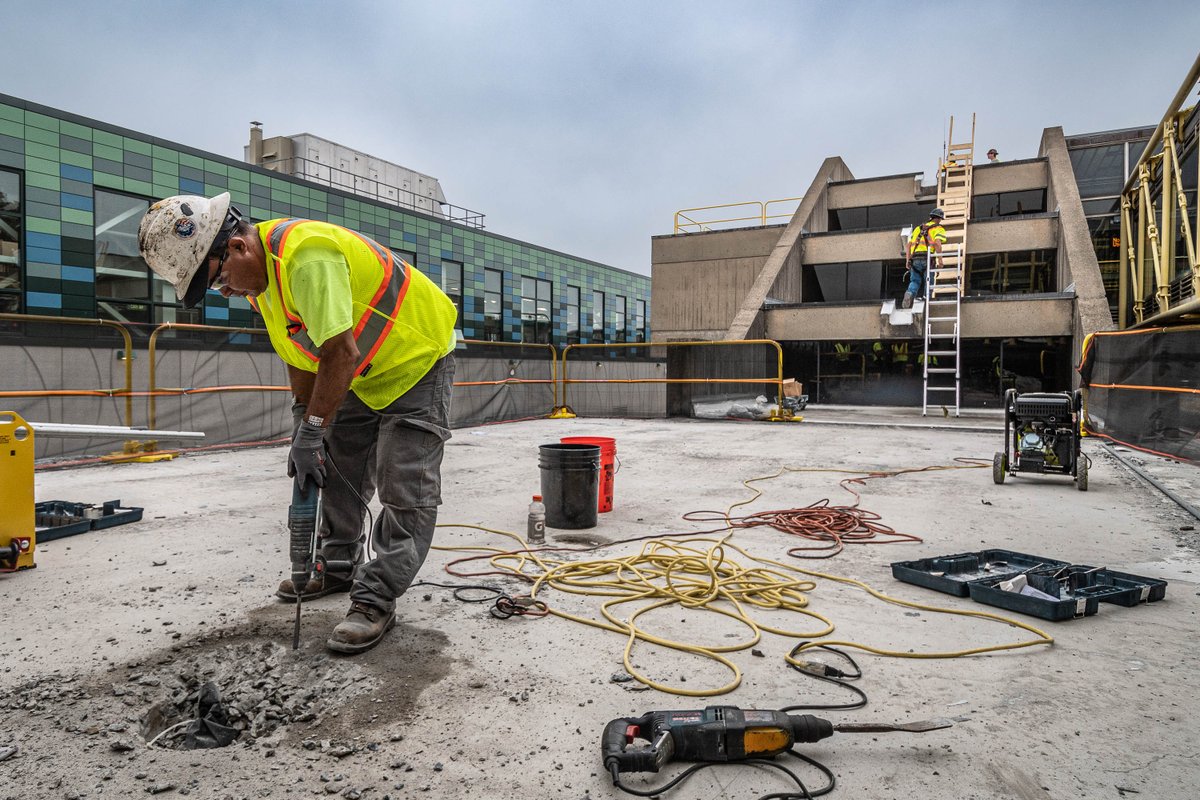 A construction worker in a yellow safety vest and hard hat chiseling at the concrete around a roof drain on the roof of Wellington station.