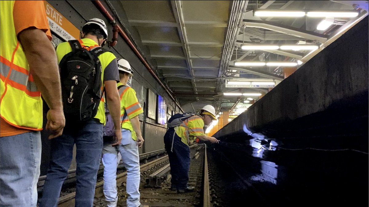 A team of inspectors in hard hats, yellow safety vests, carrying flash lights and backpacks as they walk down the Orange Line tracks at State Street station looking at wiring under the platform.
