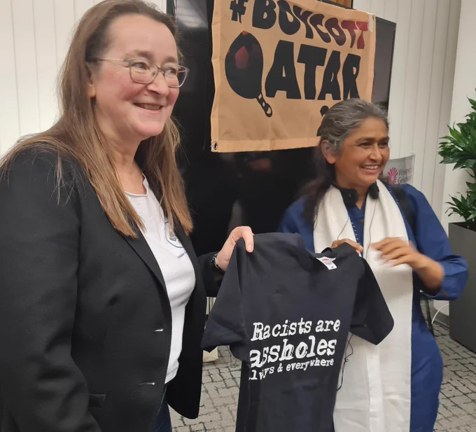 Zwei Frauen halten ein T-Shirt mit dem Aufdruck "Racists are assholes" vor dem Banner "BoycottQatar"