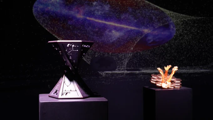 Two sculptures sit in a darkened room with visuals from Zelinskie's virtual reality art piece projected behind them. The projection shows a view of the Milky Way and a virtual version of one of the sculptures. On the right is the physical version of that sculpture, which is gold-plated and has three arms reaching out of the James Webb Space Telescope mirror. On the left in the foreground is another sculpture that looks like a twisted hourglass with equations carved into it and a hexagonal opening at top and bottom. Both pieces sit on pedestals.