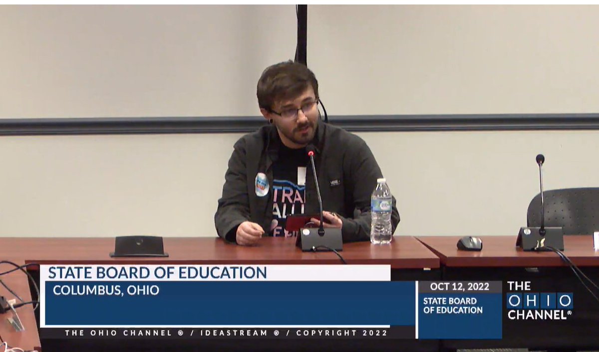 man speaking at a table, trans flag colors