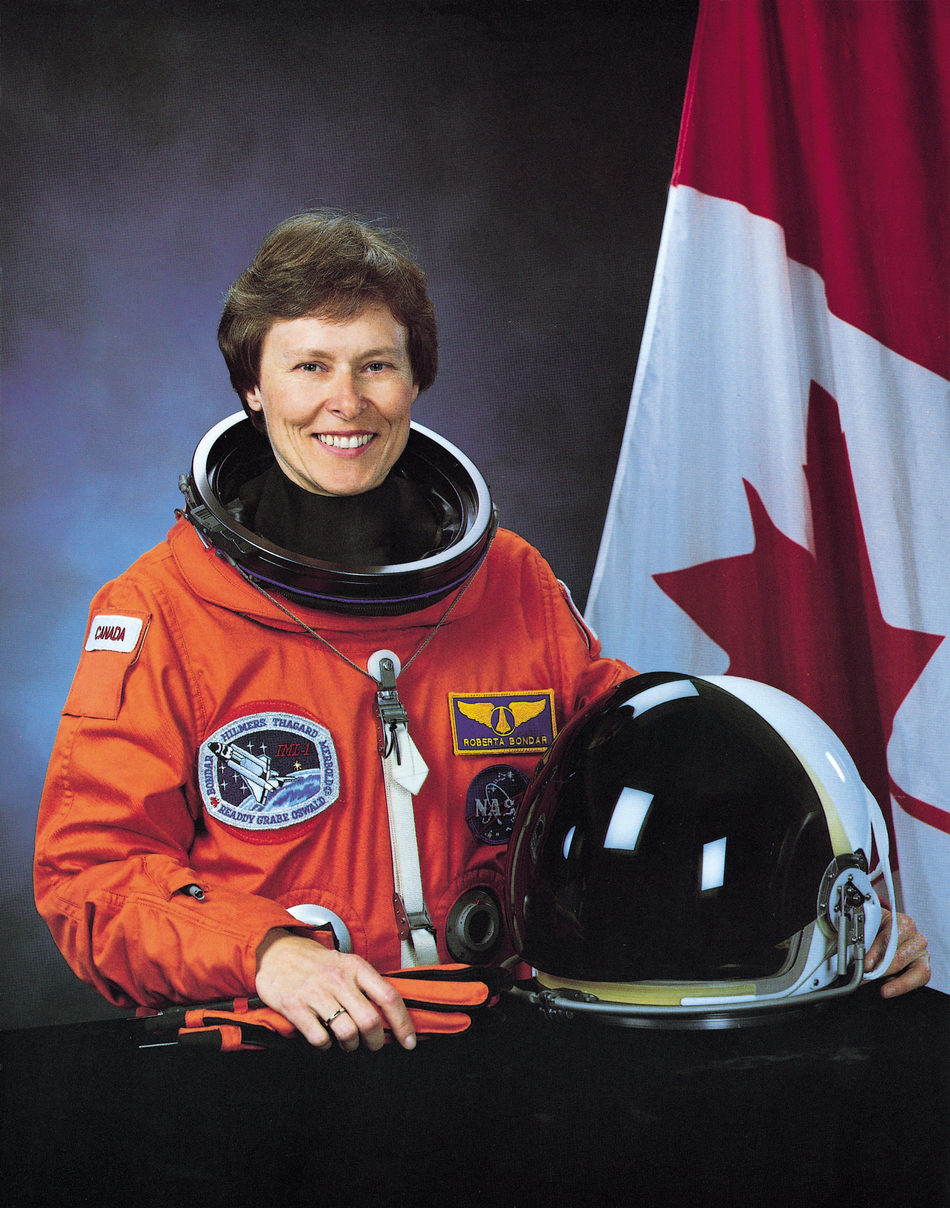 Roberta Bondar in her orange space suit beside the Canadian flag with her space helmet in hand. 