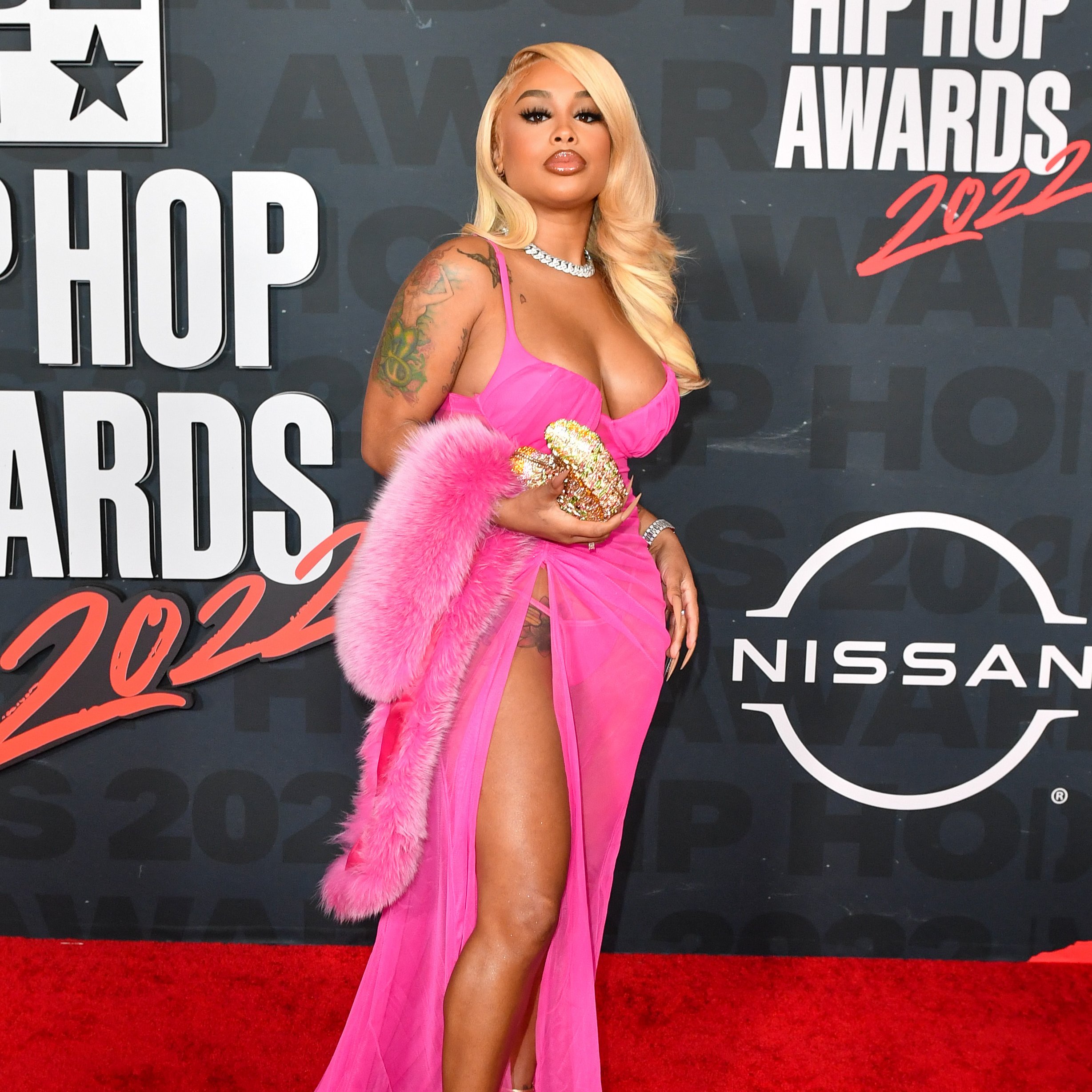 Dream Doll is rocking blonde hair with a side part. She's wearing a hot pink dress with a thigh-high slit. She fur shall is hot pink, her clutch is gold, and her necklace is diamond studded. 