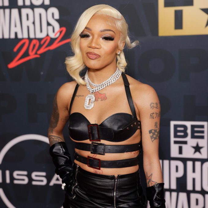 Glo is wearing a black leather dress and her hair is platinum blonde with a side part