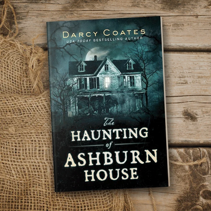 paperback copy of the haunting of ashburn house (blue and grey house in the woods under full moon) on wooden table with burlap cloth