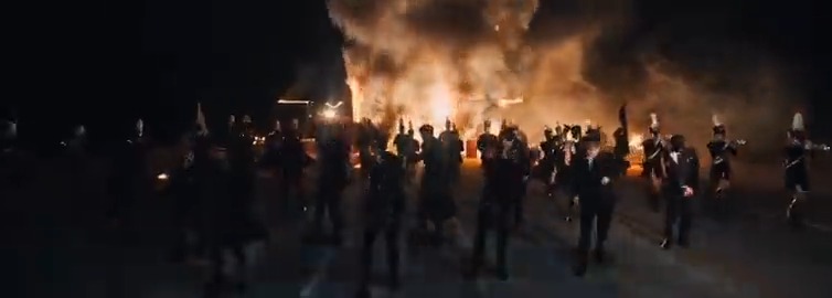 Silhouetted figures march at night in front of a large, raging fire that illuminates the dark scene.