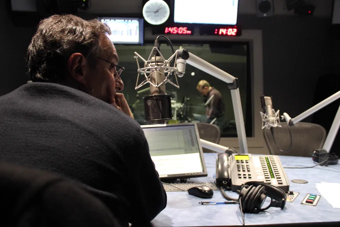 An older man with glasses, Ira Flatow, sits in a radio booth next to his mic, headphones, and the phone. In the background is a radio clock and show director.
