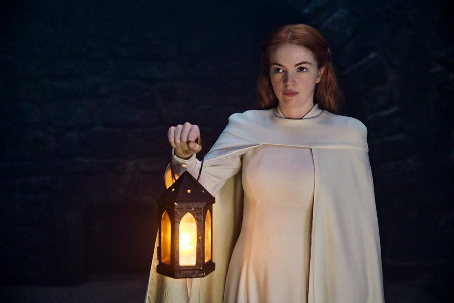 Ceara Coveney as Elayne Trakand stands in a dark stone room. She is dressed in all white, holding a lantern.