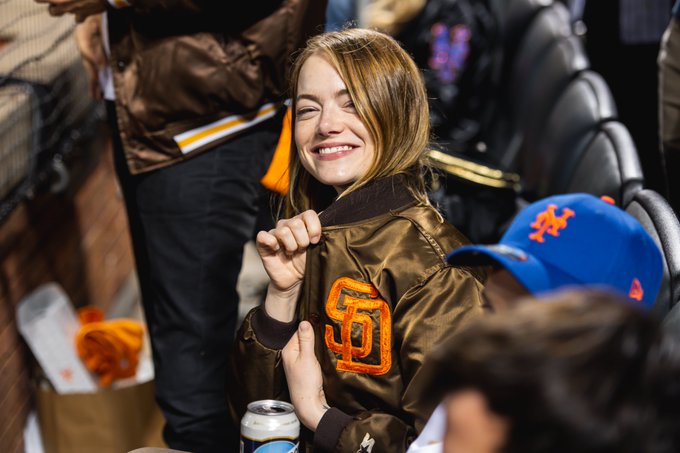 Emma Stone wearing a brown Padres jacket, smiles at the camera.