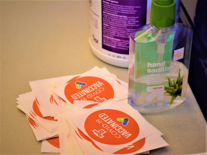 A stack of orange COVID-19 vaccination stickers from Franciscan Health next to a bottle of hand sanitizer and cleaning wipes.