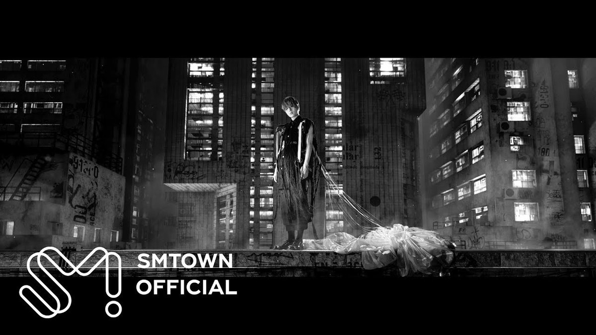 A black-and-white SMTOWN official image of a figure standing on a rooftop ledge in a dark, urban setting with illuminated apartment buildings in the background.