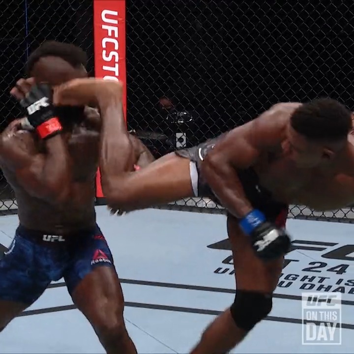 A UFC fighter on the right delivers a devastating high kick to the head of his opponent on the left, who is seen absorbing the blow with his guard raised. The cage banner reads "UFCSTORE" and the mat displays "UFC FIGHT IS ABU DHABI 24," with a "UFC On This Day" graphic visible in the corner, capturing what is being celebrated as a legendary knockout moment.