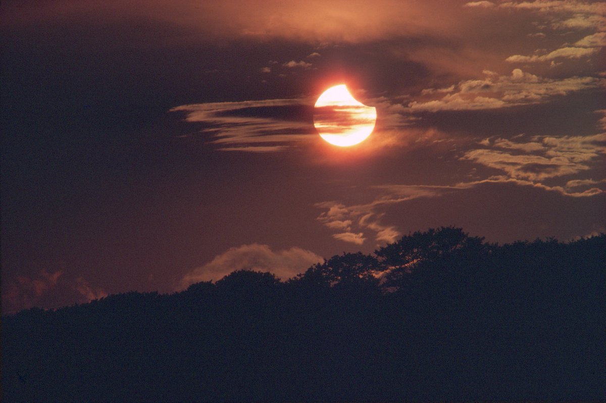 A partial solar ecipse taking place. The Sun, covered by sparse cloud, appears to have a chunk taken out of it. The sky is dark and appears a rust colour.