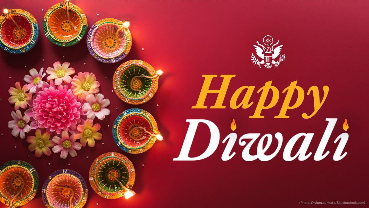 Graphic with bright cranberry red background with traditional Diwali lamps in a circular formation and flowers in the center. Text says in yellow and white, "Happy Diwali" with a white State Department eagle seal.
