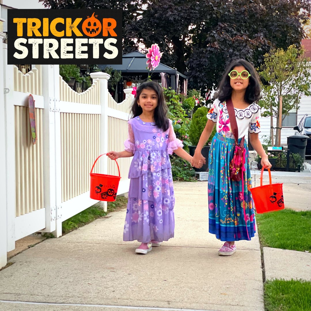 Two little girls wearing colorful and floral dresses walk on a city sidewalk while carrying orange buckets. The trick or streets logo where the o in or is a jack-o-lantern. 