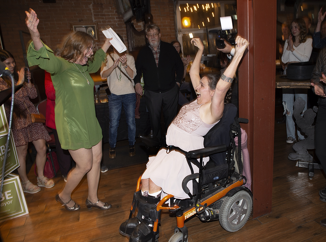 Two women raise their arms in celebration in the middle of a crowded room. One woman is wearing a green dress and standing while the other is wearing a pink dress and sitting in an orange wheelchair. They are looking at each other and yelling while smiling.