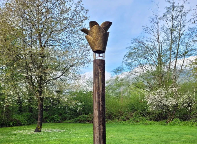 A photograph of Cedar Cone Rain Catcher on a sunny day. The cedar log with gold rain catcher stands among green grass, and flowering trees.