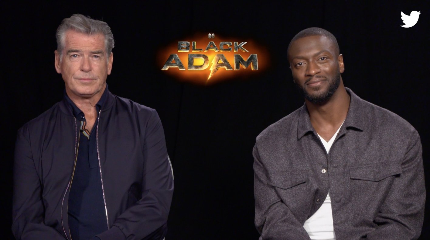 Pierce Brosnan and Aldis Hodge in front of a Black Adam logo
