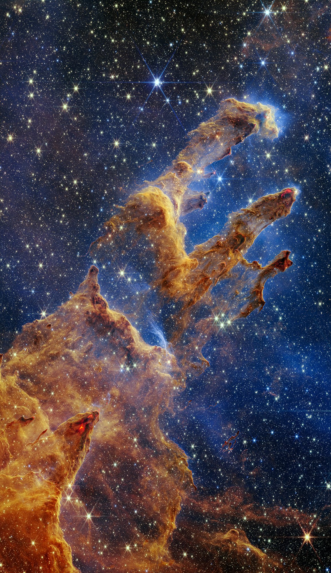 This Webb image of the “Pillars of Creation” has layers of semi-opaque, rusty red gas and dust that start at the bottom left and go toward the top right. There are three prominent pillars rising toward the top right. The left pillar is the largest and widest. The peaks of the second and third pillars are set off in darker shades of brown and have red outlines. Peeking through the layers of gas and dust is the background, set in shades of blue and littered with tiny yellow and blue stars. Many of the tips of the pillars appear tinged with what looks like lava. There are also tiny red dots at the edges of the pillars, which are newly born stars.