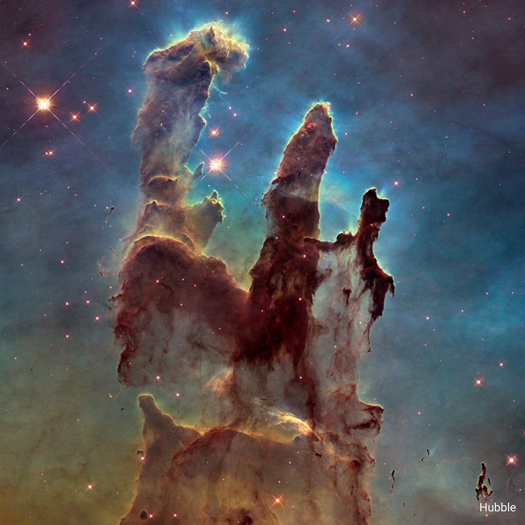 Hubble’s visible-light view of a star-forming region, the Pillars of Creation, shows darker pillars that rise from the bottom to the top of the screen, ending in three points. The background is opaque, set off in yellow and green toward the bottom and blue and purple at the top. A handful of stars of various sizes appear.