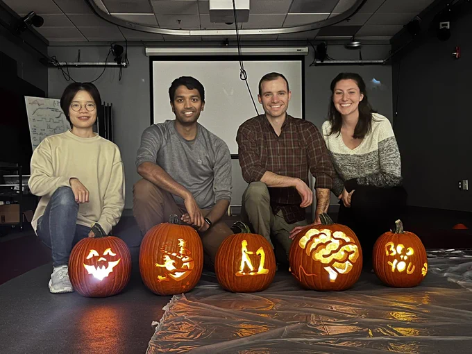 NRL lab members Ruoxi, Dheepak, Dave, and Ashley with five carved pumpkins. Pumpkins include a scary face, ghost and witch, person on a treadmill, a brain, and “Boo to you”.