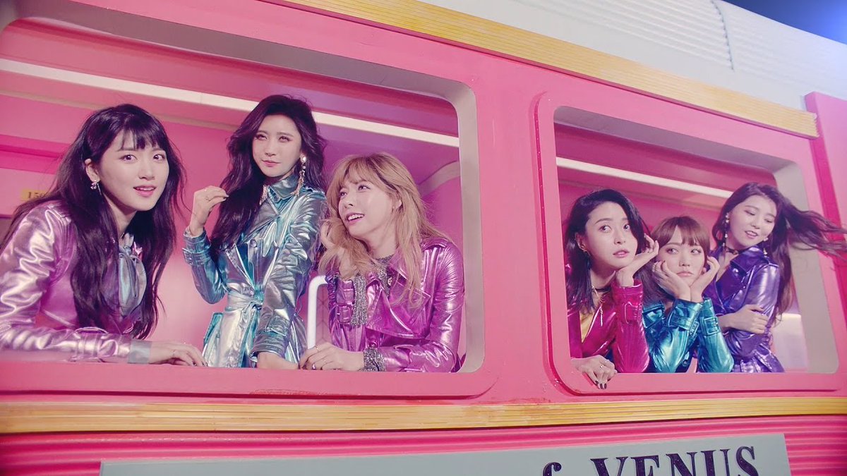 Six young women in shiny metallic purple, teal, and pink outfits look out from the windows of a bright pink vehicle or display.