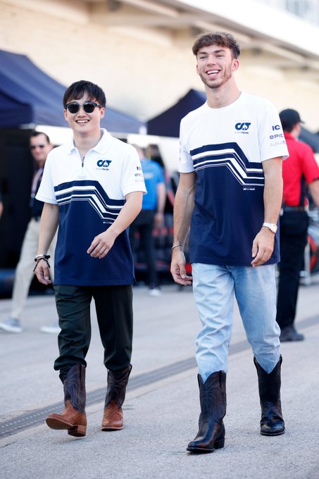 A photo of AlphaTauri drivers Yuki Tsunoda and Pierre Gasly walking through the paddock. Both are smiling from ear to ear while wearing cowboy boots. Pierre's boots are shiny and black, coming up to his shins, while Yuki's are brown at the bottom with black spots, and then black in colour from the ankles up.