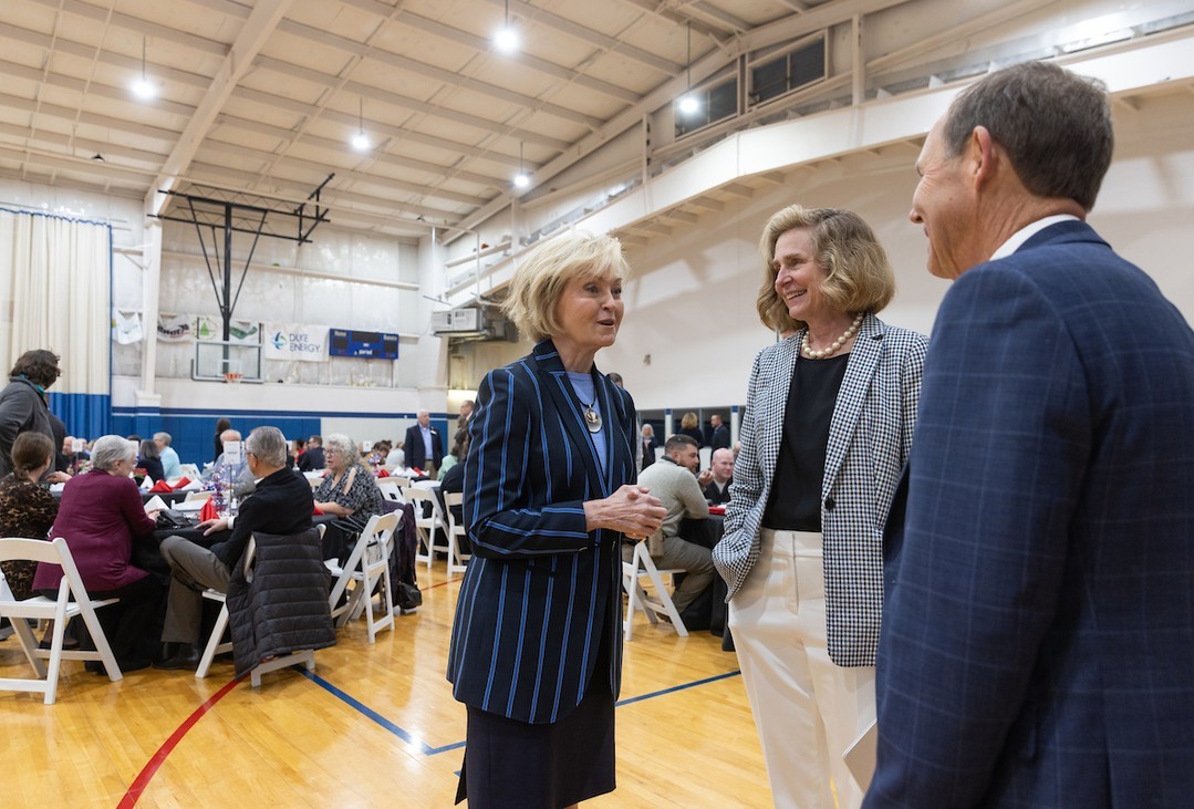 IU President Whitten stands on a basketball chatting with former Indiana Lieutenant Governor Becky Skillman and a man in a suit. Behind them people in business attire are sitting at tables eating.
