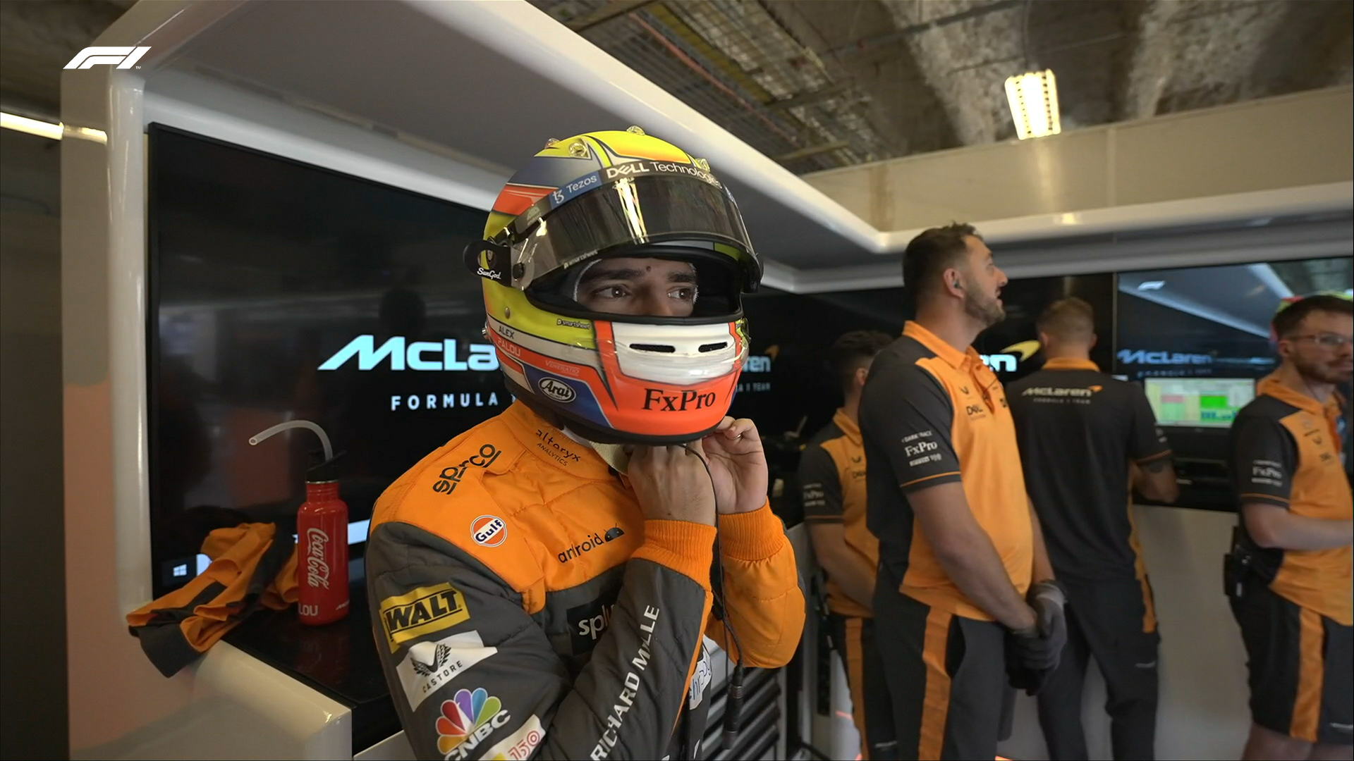 Alex Palou buckles up his helmet as he prepares for first practice in Austin with McLaren. His eyes can be seen peering through the helmet as he anticipates the laps ahead. Members of the McLaren team can be seen in the background behind him