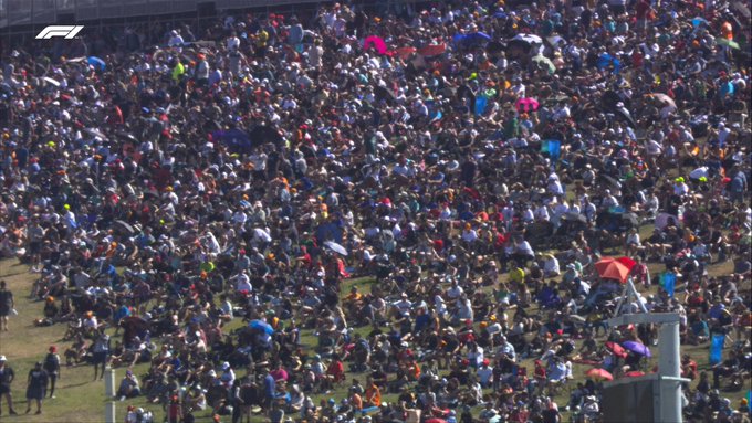 Thousands of fans can be seen strewn across the grassy embankment outside of Turn 1 at COTA, with almost none of the green grass visible on screen. The crowd is dotted with umbrellas and flags.